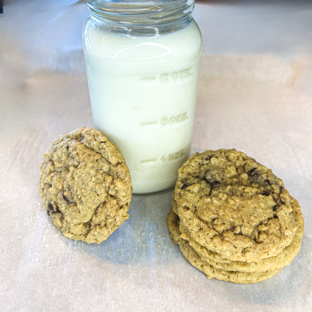 glass of farm fresh milk with cookie leaned against glass on left, on right stack of 3 cookies next to milk glass. All on top of white parchment paper.