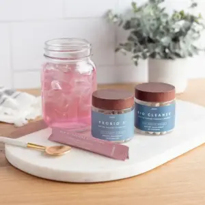 Mason glass jar with ice and light pink drink inside. bottle of probiotics with wood colored lid on clear bottle wrapped with a light blue label and text. Second bottle with same look, different contents. Contains bio cleanse. Wrapper of pink drink infront of it.